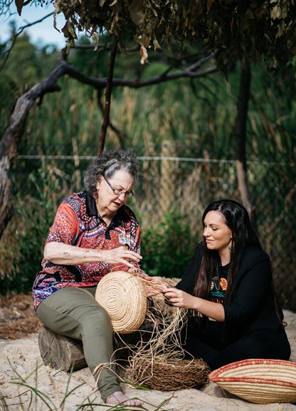 An older woman on the left is sitting on a log with a younger woman sitting in the sand weaving baskets from natural fibress