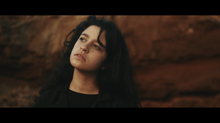 A still image from ‘Like No Other’, showing a person with long, black hair, looking wistfully into the distance. The background is out of focus.
