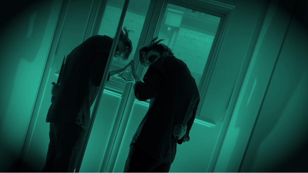 A still image from ‘Intruder’, depicting a person holding a knife behind their back and standing against a mirror. The shot is greenlit and at an angle, emphasizing shadows in the background.