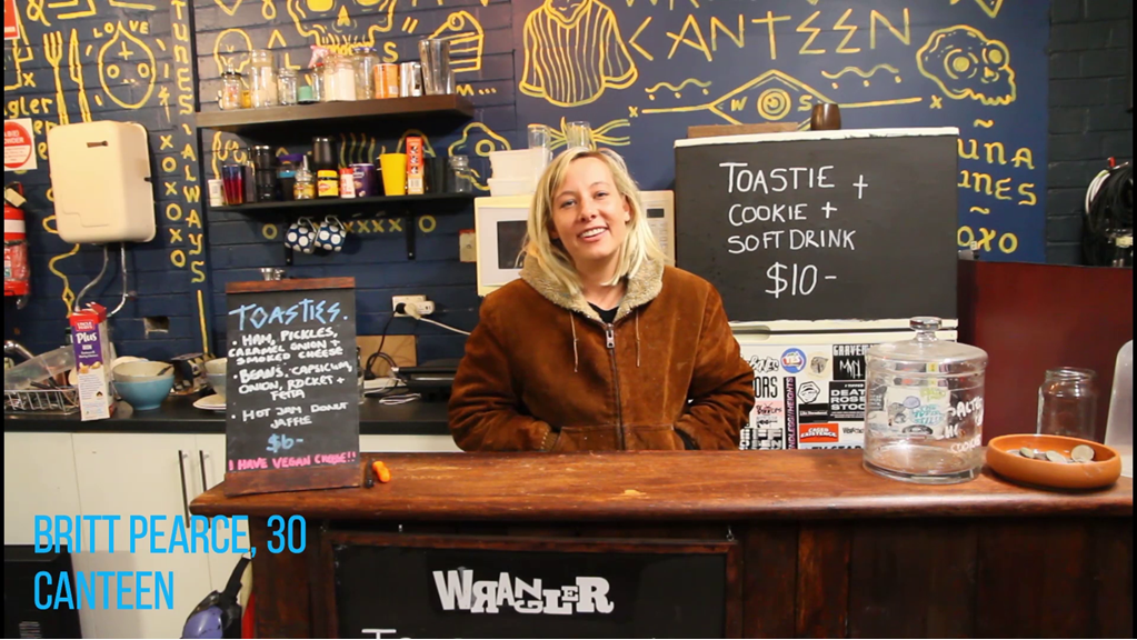 The still from ‘Wrangler’ shows a blonde haired woman sitting behind a counter of the canteen at the Wrangler venue. She is looking at the camera and smiling.