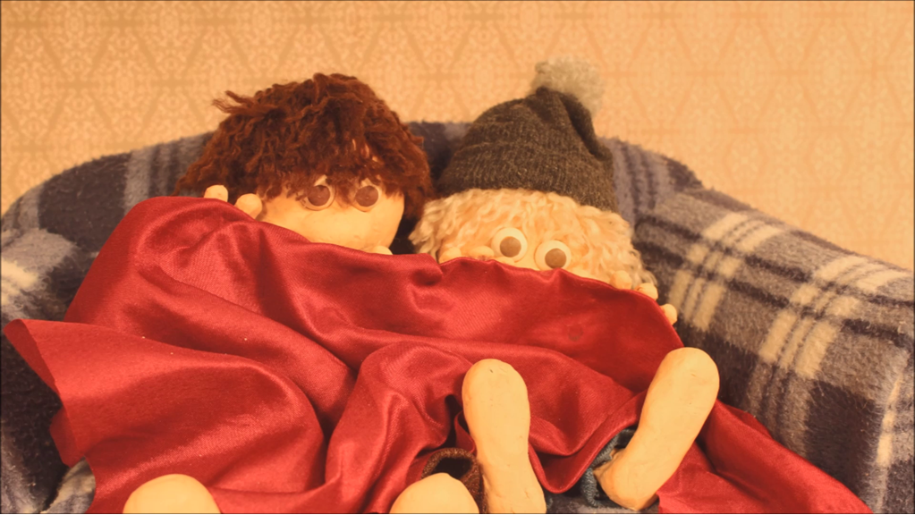 The still from the film ‘Quarantined’ shows two Claymation figures. The figures are peering out from underneath a red blanket with their feet poking out.