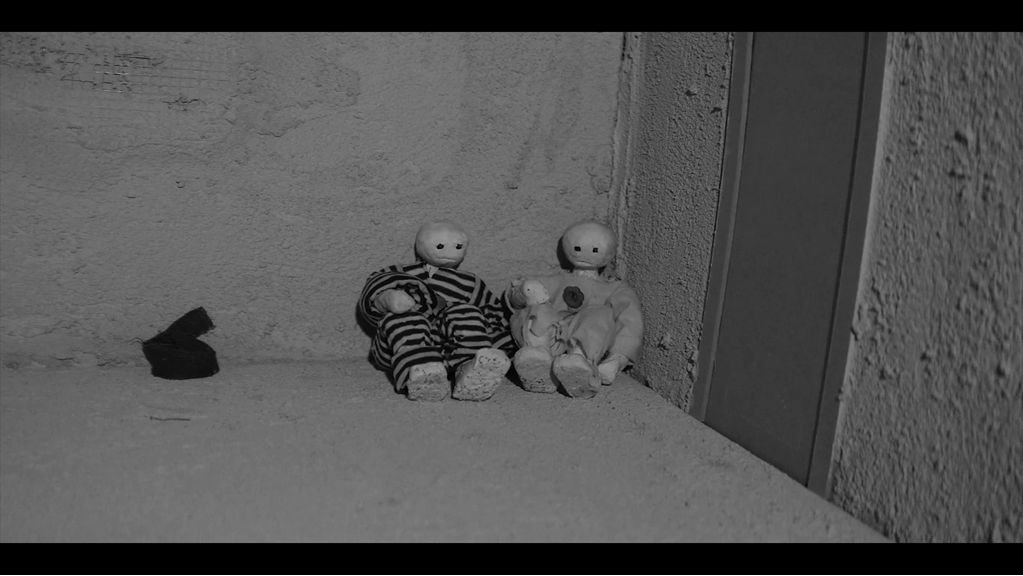 The still shows two Claymation figures sitting together in the corner of a room. One wears black and white stripes, the other a grey tunic. The still is black and white.