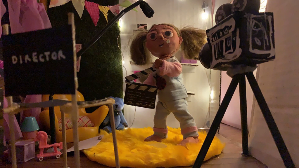 The still shows a claymation girl wearing pigtails and pajamas, holding a film clapperboard. She is standing in her bedroom, and there is a director’s chair and camera in the foreground.