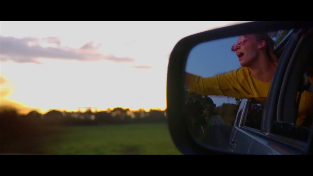 The still is a close up of a car’s side mirror. A woman is visible in the mirror’s reflection, leaning out of the car window with one arm extended. There are green fields and a brightly lit sky in the background.