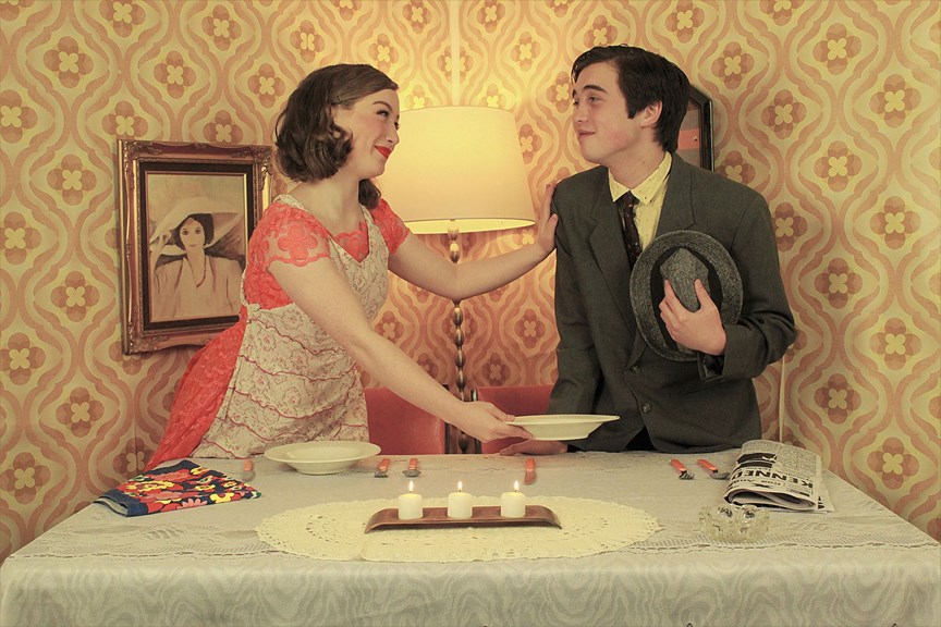 A photograph showing a woman and a man standing at a dining table in a 1960s style room. The woman is looking fondly at the man, handing him a dinner plate. 
