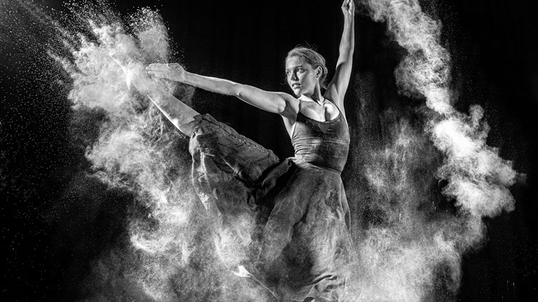 A black and white photograph depicting a dancer in motion. The dancer’s arms and legs are extended mid-leap, and a white cloud of flour surrounds them.