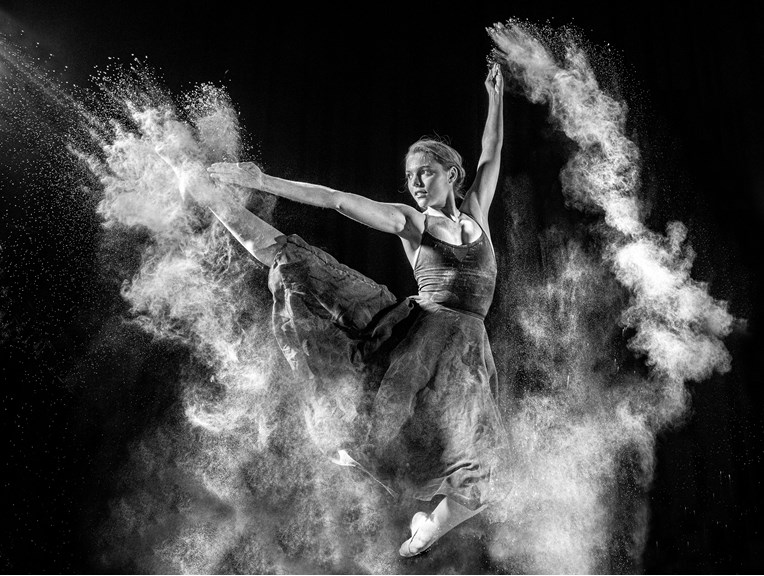 A black and white photograph depicting a dancer in motion. The dancer’s arms and legs are extended mid-leap, and a white cloud of flour surrounds them.
