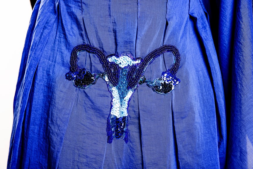 A close-up of a blue pleated dress. A uterus is beaded and sequined. It is placed to sit over the wearer’s reproductive organs.