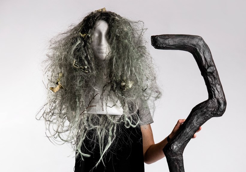 A photo of a person holding a brown staff. The staff is curved and distorted. A green and grey wig is also shown. It is long, curly, and messy with leaves through it.
