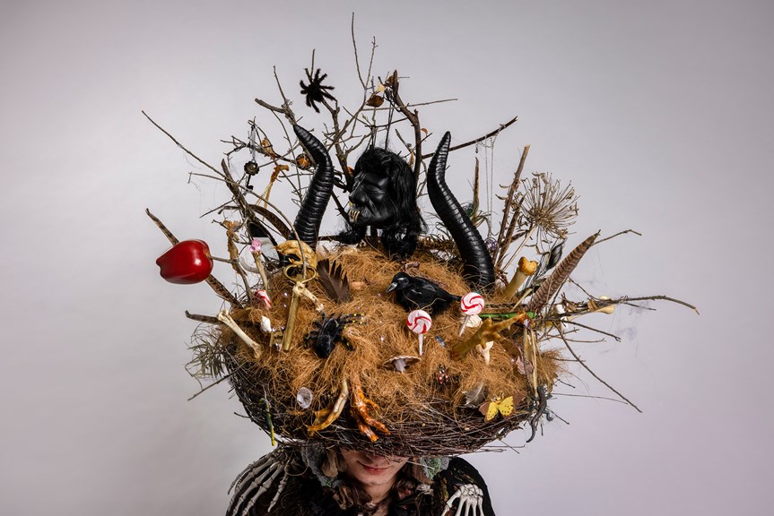 A photograph of a headpiece made to look like a nest. It is decorated with spindles, necklaces, bones, feathers, twigs, spiders, lollipops and chicken feet. A black voodoo head sits between two horns.