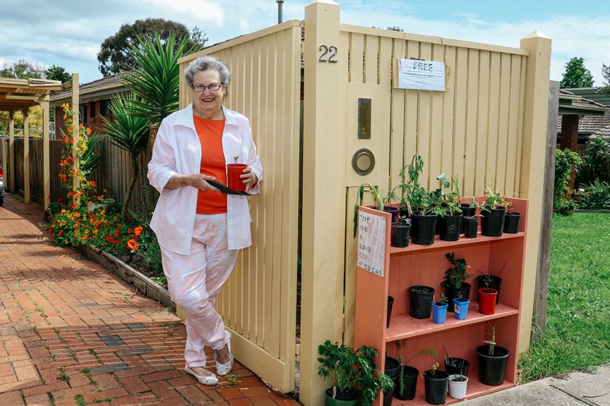 Woman dressed in white and orange holding a small pot plant at her front gate