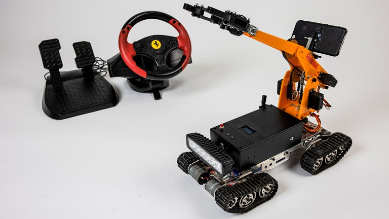 A photograph of the exploration robot. Pedals and a steering wheel are shown in the background. The orange robot is shown in the foreground with a crane and tank track wheels.