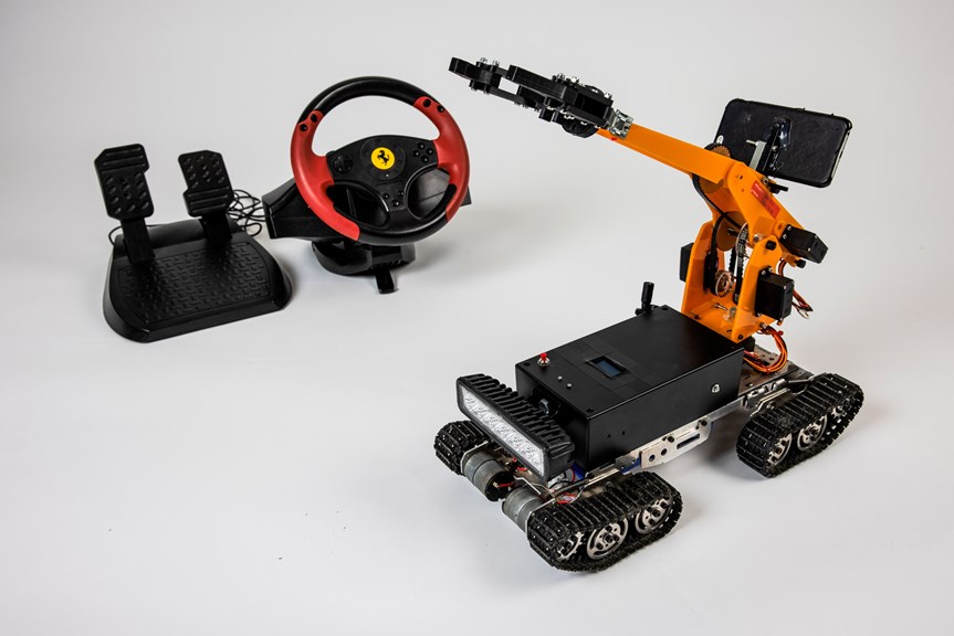 A photograph of the exploration robot. Pedals and a steering wheel are shown in the background. The orange robot is shown in the foreground with a crane and tank track wheels.