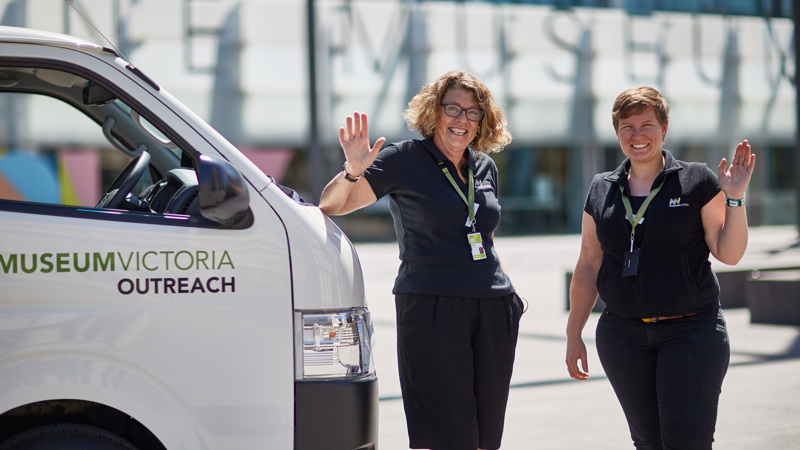 Two women wearing black are waving. They stand next to a white van outside Melbourne Museum.