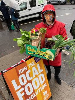 Person wearing a mask and holding a box full of produce standing next to an a-frame sign
