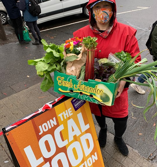 Person wearing a mask and holding a box full of produce standing next to an a-frame sign