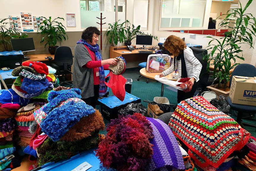 Two woman in an office sorting knitted beanies, gloves, scarves and blankets