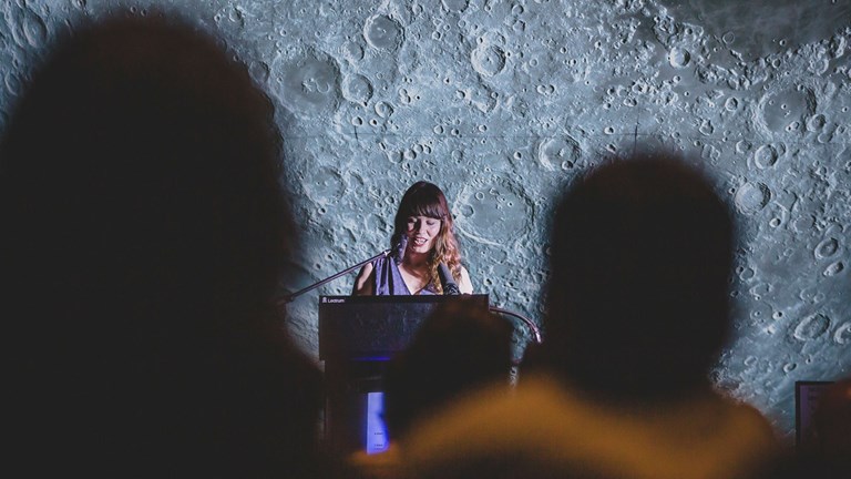 A woman stands at a lectern with a Moon background and out-of-focus audience in the foreground. 