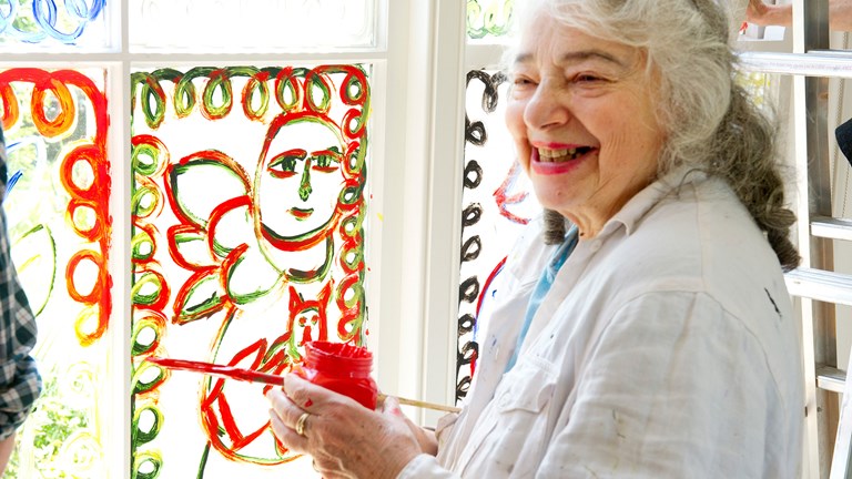 Older woman smiling holding a red paint pot and brush.