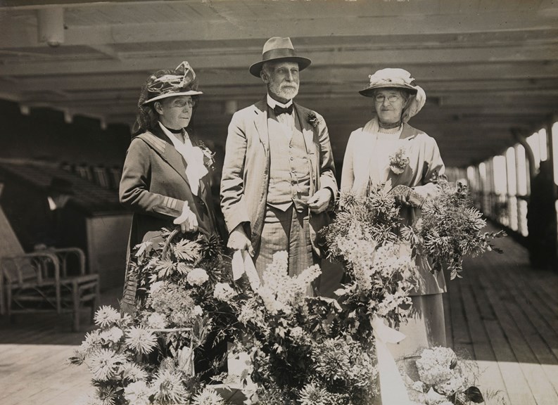 Black and white photograph of three people, two woman and man, standing on a ship with many bunches of flowers