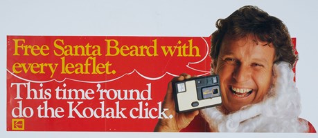 White, red and yellow poster with an image of a man with a santa beard pull below his chin holding a small camera. Text " Free Santa beard with every leaflet. This time "round do the Kodak click"