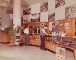 People standing in a photographic shop. Two male shop assistants stand behind the counter while two customers stand with them in front of the counter.
