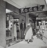 People walking along a footpath and standing in the doorway of a photographic shop.