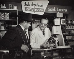 Man standing behind a counter in a photographic shop showing a photograph of a woman to another man standing on the other side of the shop counter.