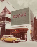 Exterior of a Kodak photographic shop. Car parked in street. People walking on footpath.