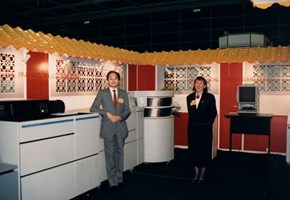 Woman and man with yellow nametags standing in a trade show/ conference stand with business imaging equipment around them.