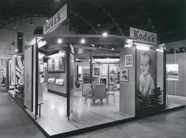 Exhibition display stand viewed from its corner showing lighting and steel roof supports above the stand, and various divided display areas.
