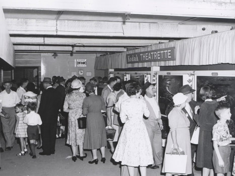 Crowd of women, men and children outside 'Kodak Theatrette' with photographs mounted in light box on right wall.