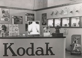 Kodak exhibition/trade show display stand with service counter, and display cases in wall. Posters and photographs decorate the display.