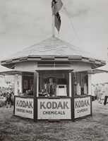 A Kodak staff member stands smiling to camera at the counter of the booth, and there are products arranged within. There is a large Kodak flag on top of the pointed roof and the windows of the booth are shuttered. Crowds and other buildings and pavilions are visible in the background.