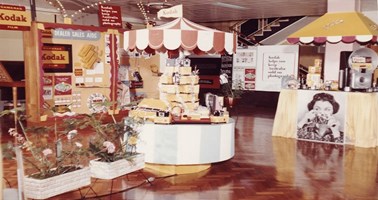 A wholesale trade display for dealers/industry in the 1960s. There is a board covered in different dealer sales aids. In the foreground are decorative pot plants and a stand filled with Kodak products. A board at the back of the room reads: 'Kodak helps you keep Australia sold on photography'.