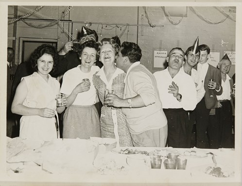 Group of men and women drinking and laughing. A man is kissing an older woman in the foreground, and behind them several men are drinking and laughing.