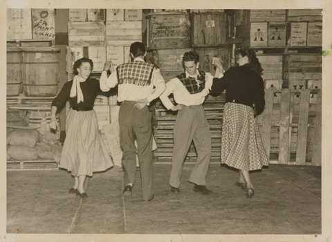 Two couples dancing in a factory storeroom, spinning in a circle holding their hands high