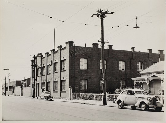 Black and white photograph of a brick building with with car parked outside in the street.