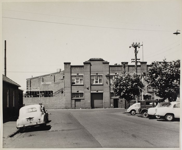 Black and white photograph of a brick building. There are cars parked in the street.