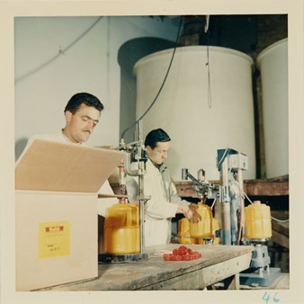 Two male workers in white overalls with yellow plastic containers underneath hoses and taps. Two large vats in background and box in foreground.