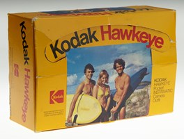Yellow camera box with a photograph of two men and woman wearing swimwear outdoors. The two men are holding surf boards. Black and read text "Kodak Hawkeye"
