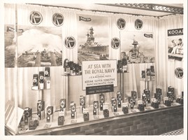 Display window with folding cameras mounted on plinths and along the floor with a central text panel reading: 'AT SEA WITH / THE ROYAL NAVY' and three large photographic prints along the back wall of naval ships. 