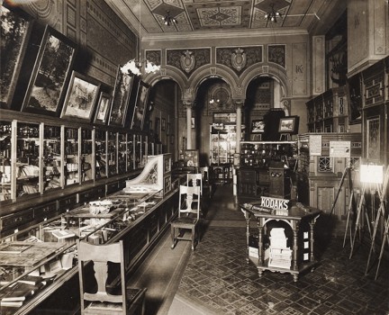 A shop interior, display counters and cases. Photographs hanging on walls. Chairs for clients to be served in. There are columns at the rear of the store with elaborate paint and plasterwork above them. The letter 'K' is painted in the centre of wall and ceiling friezes.
