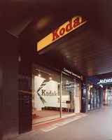 Kodak shop entrance with glass door and frosted Kodak 'K' logo on the door. There is a Kodak sign hanging from the awning and other shops visible along the footpath in the background.
