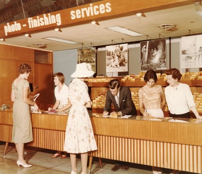 Five women and one man in a shop that sells photographic materials. Three of the women and the man are behind the shop counter. Two women are standing in front of the counter.