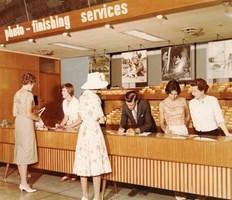 Five women and one man in a shop that sells photographic materials. Three of the women and the man are behind the shop counter. Two women are standing in front of the counter.