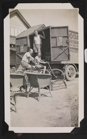 Man and a child riding a bicycle with a man standing in the back of an open delivery van behind them.