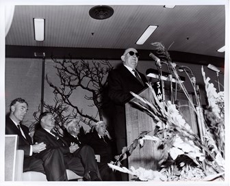 Man standing a a lectern speaking. Other men are seated behind him.