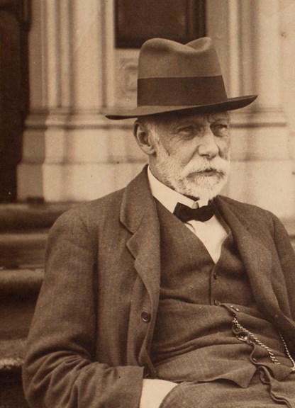 A bearded, seated man in a hat and bow tie