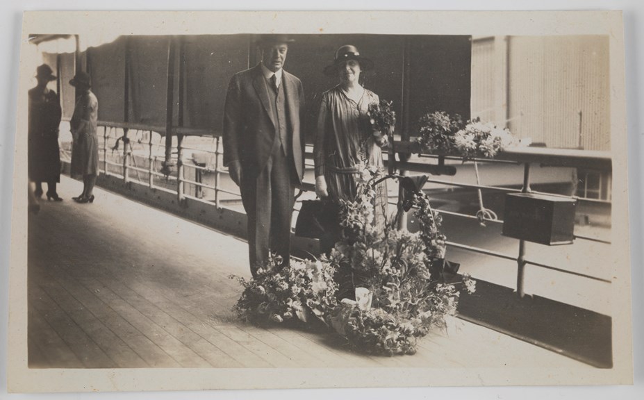 Man and woman standing on deck of ship, surrounded by floral arrangements. There are women standing in the background.
